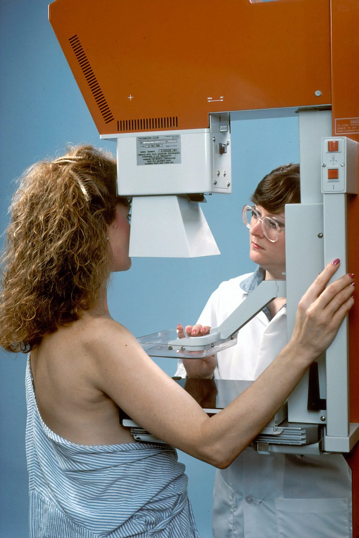 A woman undergoing a traditional mammogram, positioned at a fixed imaging unit by a clinician